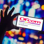 The Ofcom logo displayed on a smartphone behind held by someone, with bright coloured lights in the background