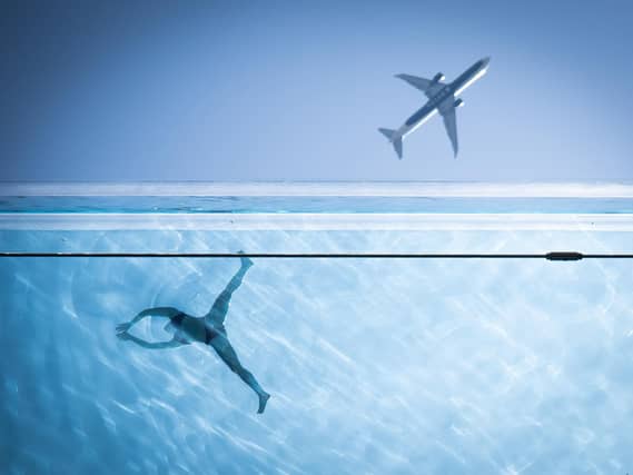 A woman swims in a clear pool while above an airliner flies past, both viewed from below