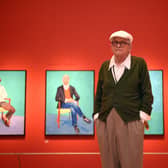 David Hockney with some of his portraits behind him hung on a red wall