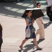 A woman walks across paving in the sunshine sheltering under a black umbrella