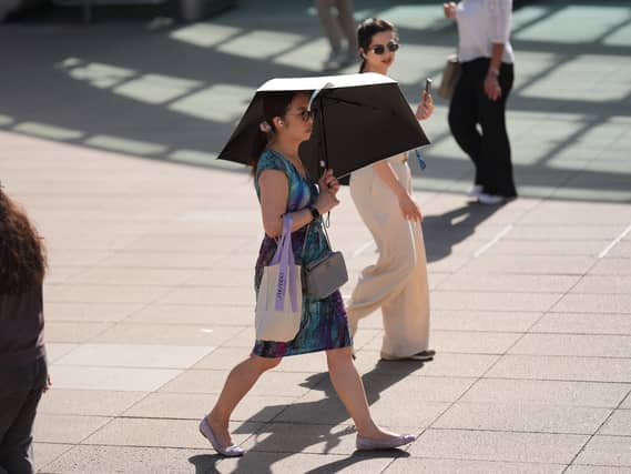 A woman walks across paving in the sunshine sheltering under a black umbrella