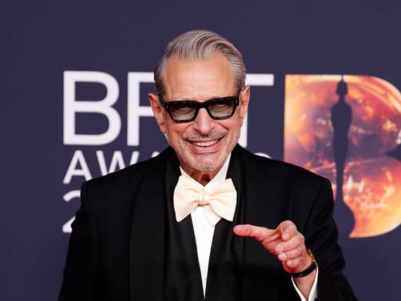 Jeff Goldblum smiling with his left hand raised and pointing wearing a black suit and white bow tie at the Brit Awards