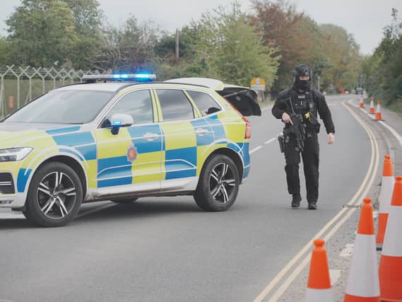 An armed policeman walking on a road, next to a police car with its blue lights on