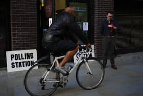 A Labour Party teller wearing a red rosette standing outside the N1C Centre polling station in London as a cyclist rides past