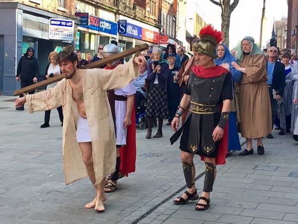 Dressed in only a cream robe and white shorts, a man portraying Jesus walks his cross down a busy highstreet alongside two Roman soldiers and followed by a crowd of actors and audience as part of the Shrewsbury Passion Play 2019