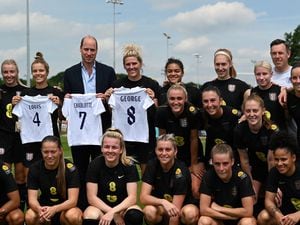 The Duke of Cambridge holding a England football jersey bearing the names of his three children and with the England women football team during a visit to St George's Park ahead of their game at Molineux